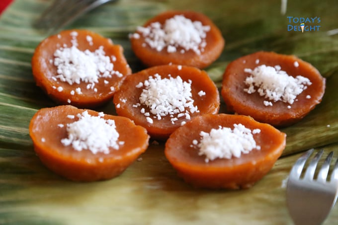 Six sticky brown rice cakes topped with grated coconut on banana leaves with a fork are Filipino sweets known as kutsinta or kutchinta.