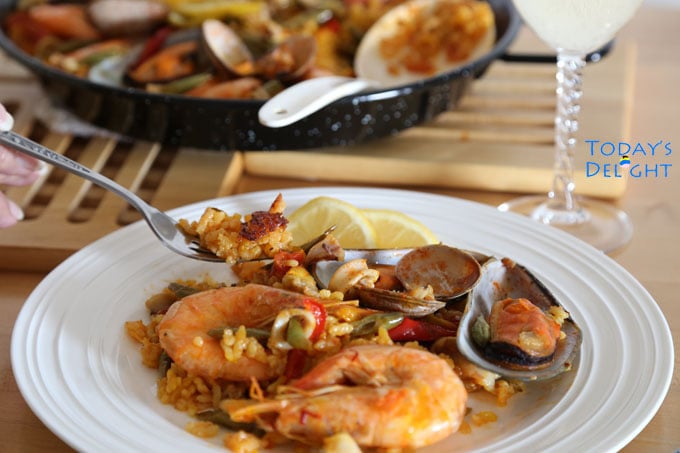 Plated seafood paella with shrimp, manila clam, mussel, lemon and rice on a fork while in the background is the paella pan and a drink.
