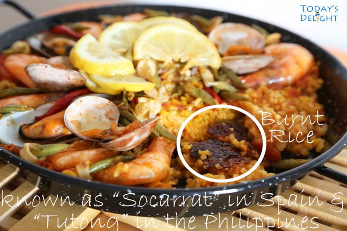 Seafood paella cooked on a paellera topped with mussels, manila clams, sliced squid, green beans and slices of lemon in the middle. It is showing socarrat, burnt rice or crust at the bottom of pan.