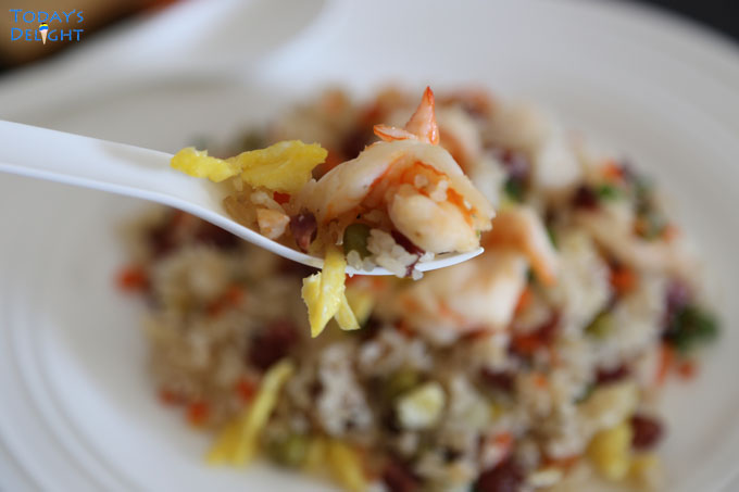Fried rice with shrimp and scrambled eggs on a spoon and a blurred plate of yang chow fried rice.