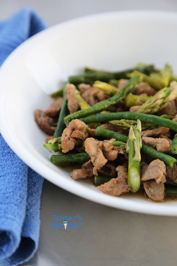 Stir fried green beans with pork and asparagus on a serving dish and blue napkin on the side.