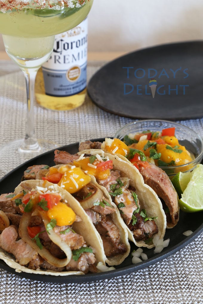 Four pork tacos topped with mango salsa and a side of mango salsa and lime wedges on a sizzling plate. Spicy margarita, corona beer and an empty sizzling plate in the background.