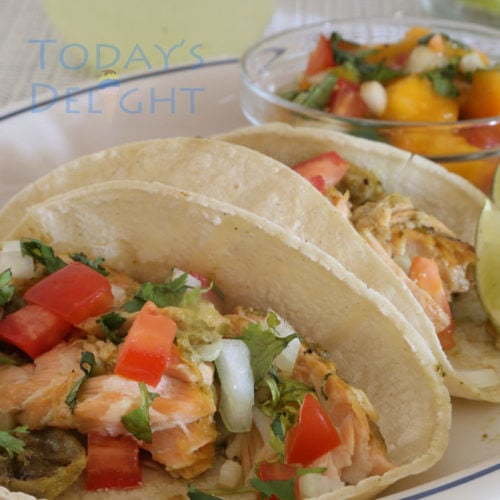 Two salmon fish tacos on a plate with lemon wedges and mango salsa.