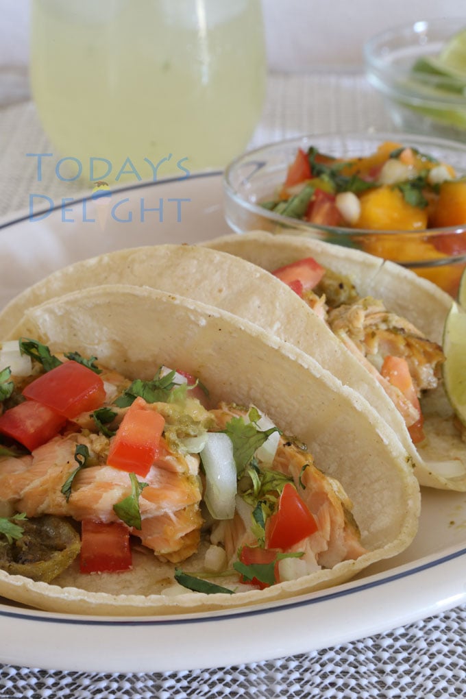 Two salmon fish tacos on a plate with lemon wedges and mango salsa.