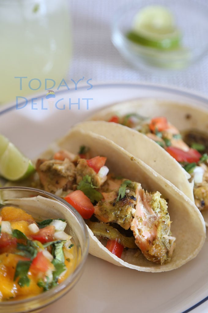 Two salmon fish tacos on a plate with lemon wedges and mango salsa. A glass of margarita and lime wedges on the side.