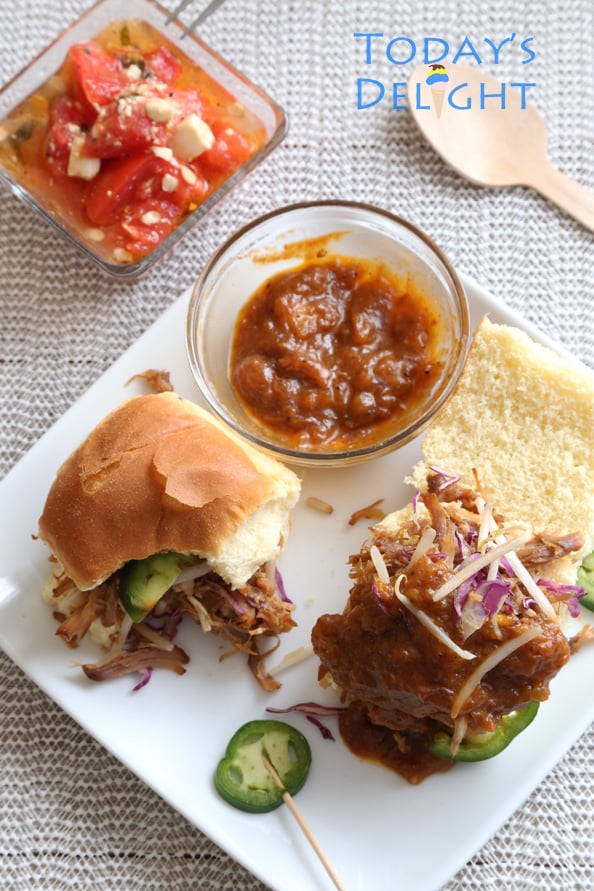 A plate of two pieces sweet and spicy pulled pork sliders with jalapenos, a side of sauce and watermelon salad. One slider with a bite and sauce is added to one.