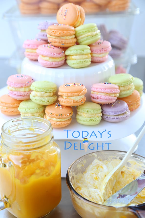 Three layers of colorful French macarons and more in containers probably about 50 pieces total. A jar of mango jam and a bowl of macaron filling.