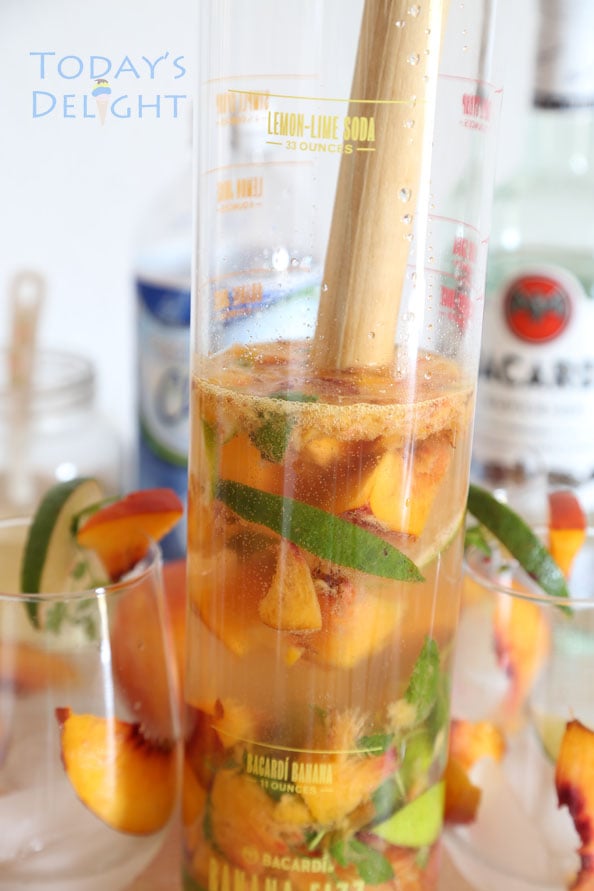 Nectarine mojitos ingredients muddled in a tall glass, 2 garnished empty glasses, rum and club soda in the background.