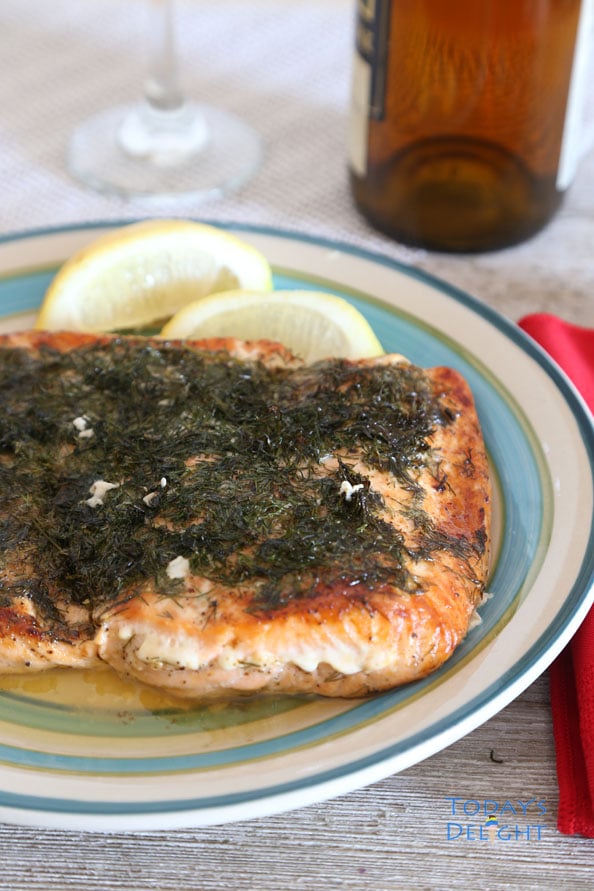 Baked salmon topped with dill and
two lemon wedges on a plate with a. salmon on a fork. A glass, wine bottle and napkin in the background.
