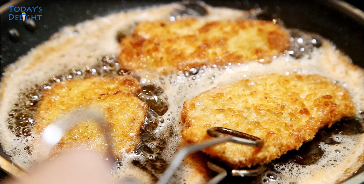 Three fried breaded pork chops in a pan are done and removed from frying pan with a tong.