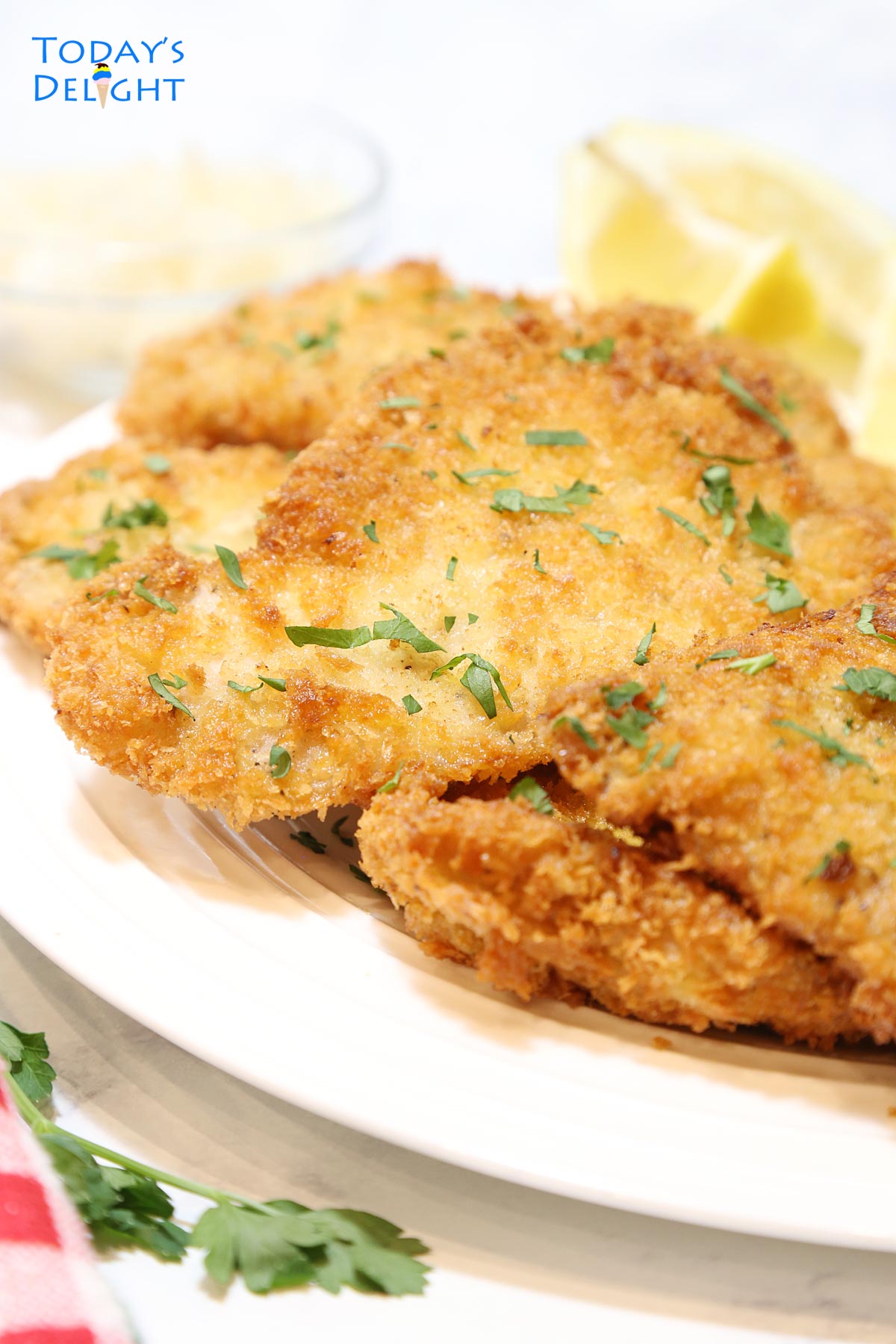 Fried pork cutlets on a plate garnished with chopped parsley and lemon wedges and more lemon on the side.