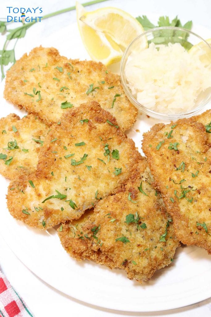 Pan fried breaded pork loin cutlets on a platter with a side of sauerkraut and lemon wedges.