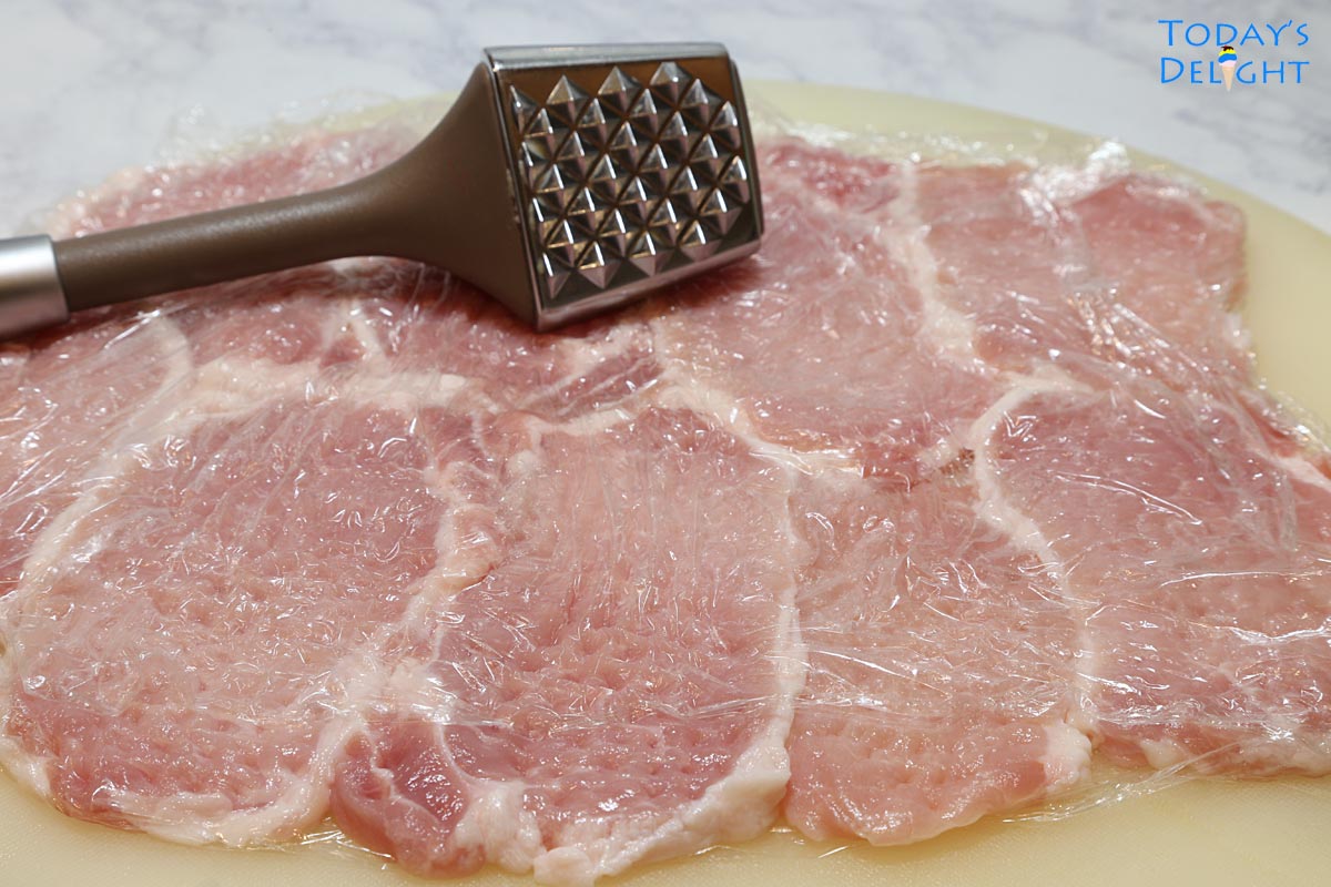 Sliced cutlets of pork tenderloin on cutting board covered with plastic wrap and pounded with a meat mallet to thin the pork.