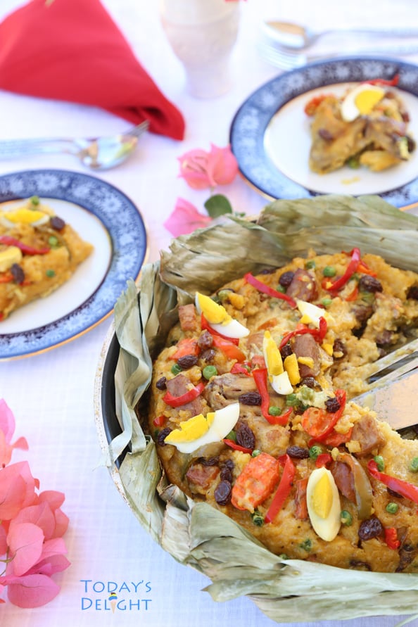 Bringhe (Philippines native paella) made of glutinous rice topped with meat, hard-boiled eggs, strips of red bell pepper, raisins and green peas on a pan lined with banana leaves served in two plates. Table has vase, flowers and utensils.