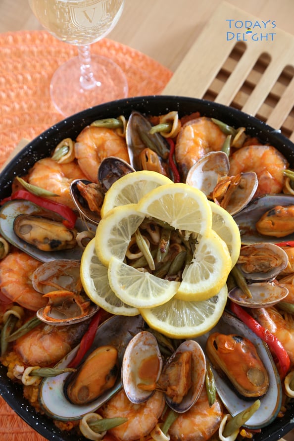 Seafood paella covered with shrimp, mussels, clams, squid, green beans, strips of red bell pepper and at the center are sliced lemon on a paella pan.