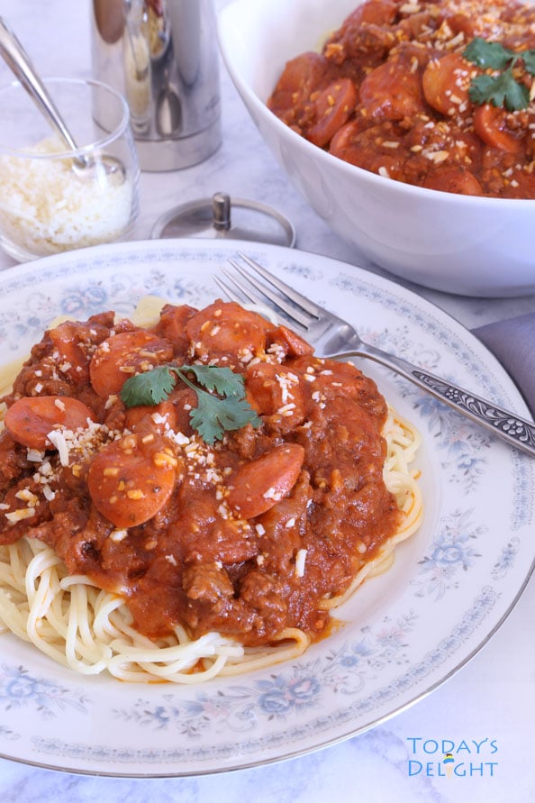Filipino style spaghetti with hot dog in meat sauce in a serving bowl and on a plate with a side of parmesan cheese.