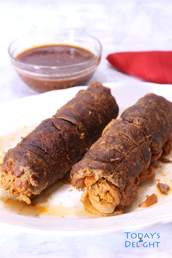 Two cooked beef morcon is a marinated beef roulade on a platter, a sauce on the side and a red napkin.