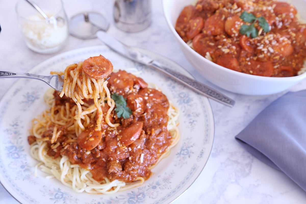 Pinoy style spaghetti with hot dog in meat sauce in a serving bowl and on a plate with a side of parmesan cheese.