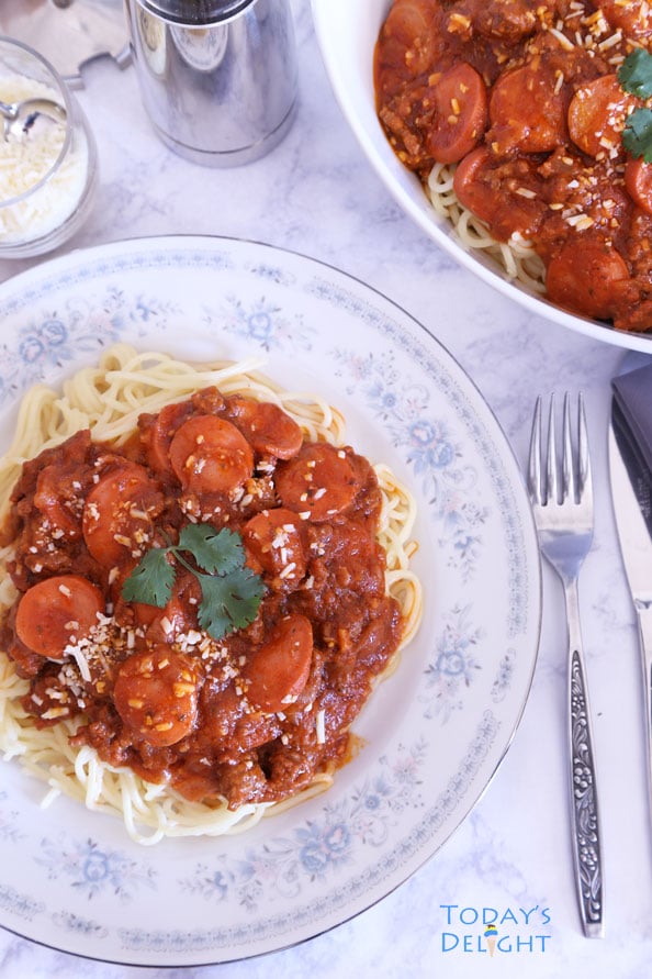 Spaghetti with hot dog in meat sauce in a serving bowl and on a plate with a side of parmesan cheese.