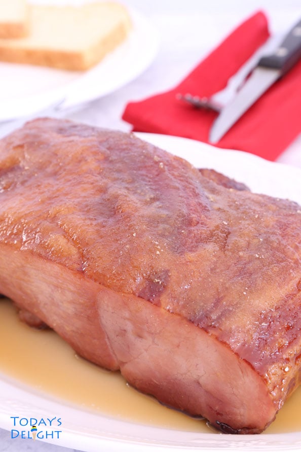 Cooked pineapple carve ham with brown sugar glaze on a platter, two pieces of bread on a plate and a knife and fork on top of a red napkin.