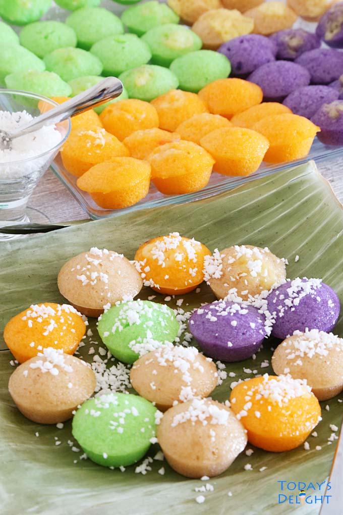 Colored bite-sized rice cakes known as puto calasiao,on a banana leaves and tray. Some rice cakes are topped with grated coconut. Grated coconut with serving spoon on the side.