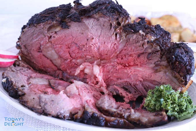 Medium cooked bone-in rib eye roast with kale garnish and baked baby potatoes in the background.