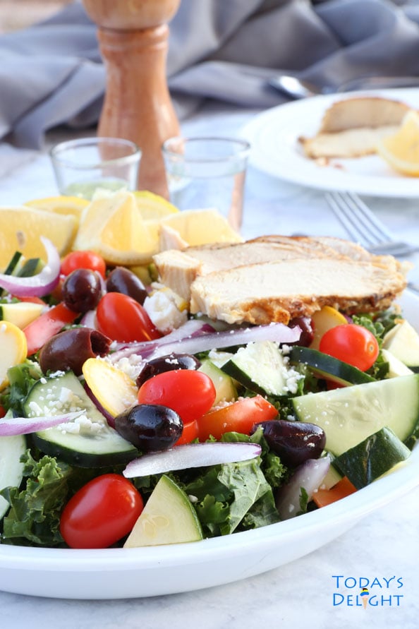 Mediterranean Kale Salad with Kalamata Olives, grape tomatoes, red onions, and Feta on a plate with lemon wedges and lemon-pepper chicken.