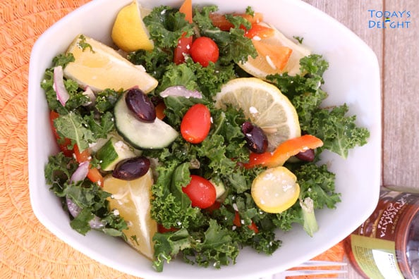 Mediterranean Kale Salad with Kalamata Olives, grape tomatoes, red onions, feta crumbles, and lemon wedges on a salad plate.