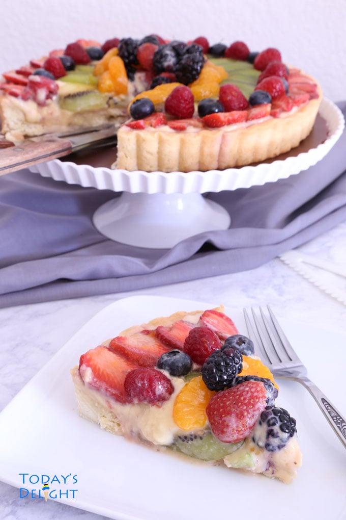 A sliced of fruit tart with fork on a plate and the whole fruit tart on a cake stand in the background.