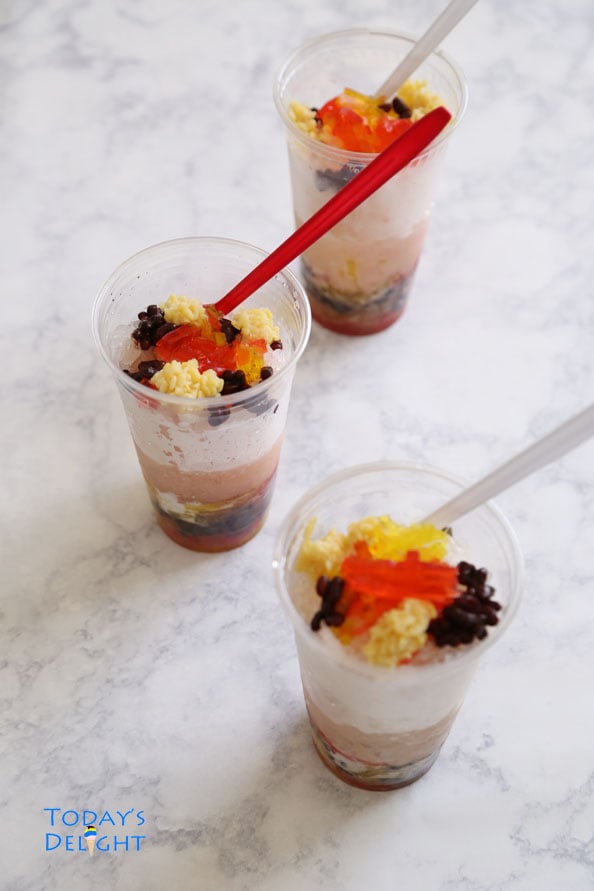 Three Asian desserts with mung beans, red beans, agar-agar, coconut milk, and shaved ice in plastic cups with red spoons.