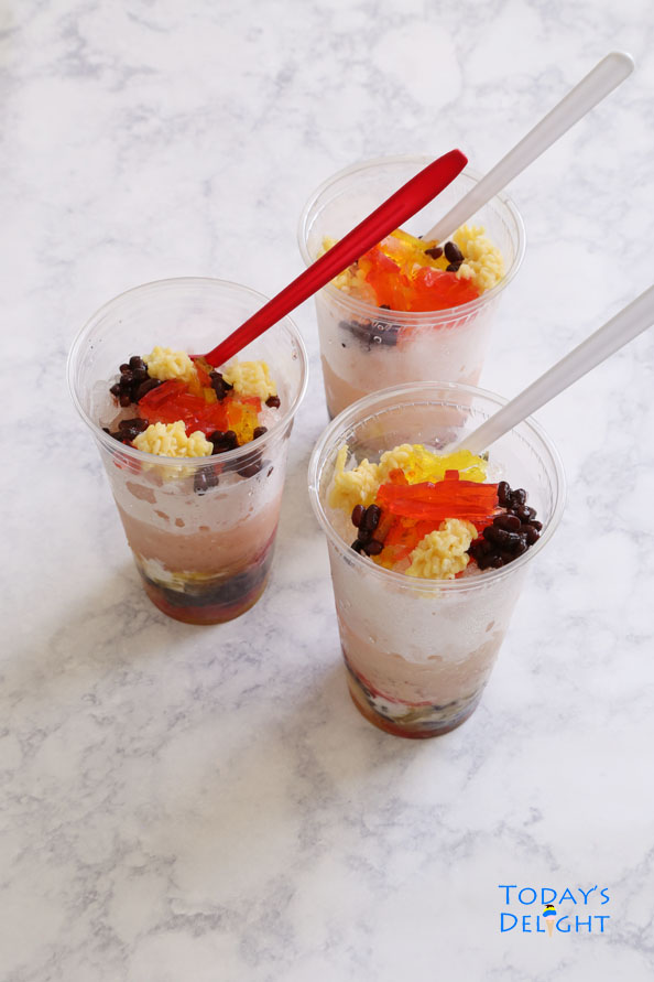 Three Asian desserts with mung beans, red beans, agar-agar, coconut milk, and shaved ice in plastic cups with red spoons.