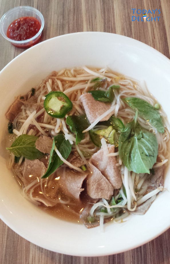 A big serving of Vietnamese beef pho consisting of beef broth, rice noodles, meat and garnished with mint leaves, jalapeno and bean sprouts and a side of sriracha.