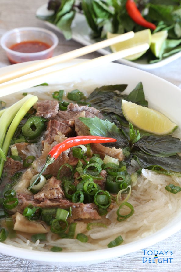 A big serving of Vietnamese beef pho consisting of beef broth, rice noodles, herbs, and meat, and garnished with bean sprouts, Thai basil, jalapeno, green onions, chili peppers, and wedge of lime. Chopstick on the bowl, serving of sriracha and more garnishes on the side.