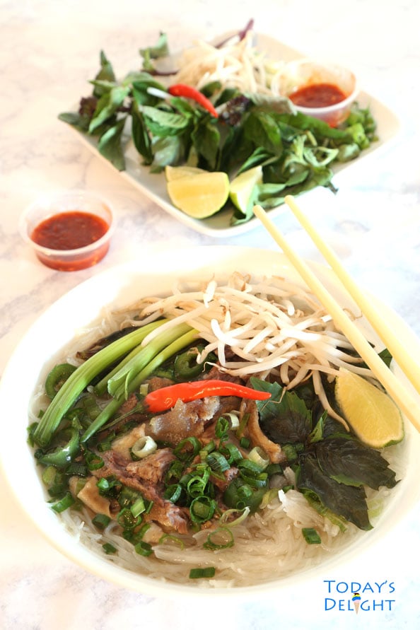 A big serving of Vietnamese beef pho consisting of beef broth, rice noodles, herbs, and meat, and garnished with bean sprouts, Thai basil, jalapeno, green onions, chili peppers, and wedge of lime. Chopstick on the bowl, serving of sriracha and more garnishes on the side. 