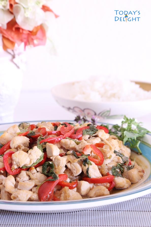 Basil Chicken flavored with basil, Thai chili, jalapeno and red bell pepper on the serving platter. A bowl of white rice and a a vase on the table too.