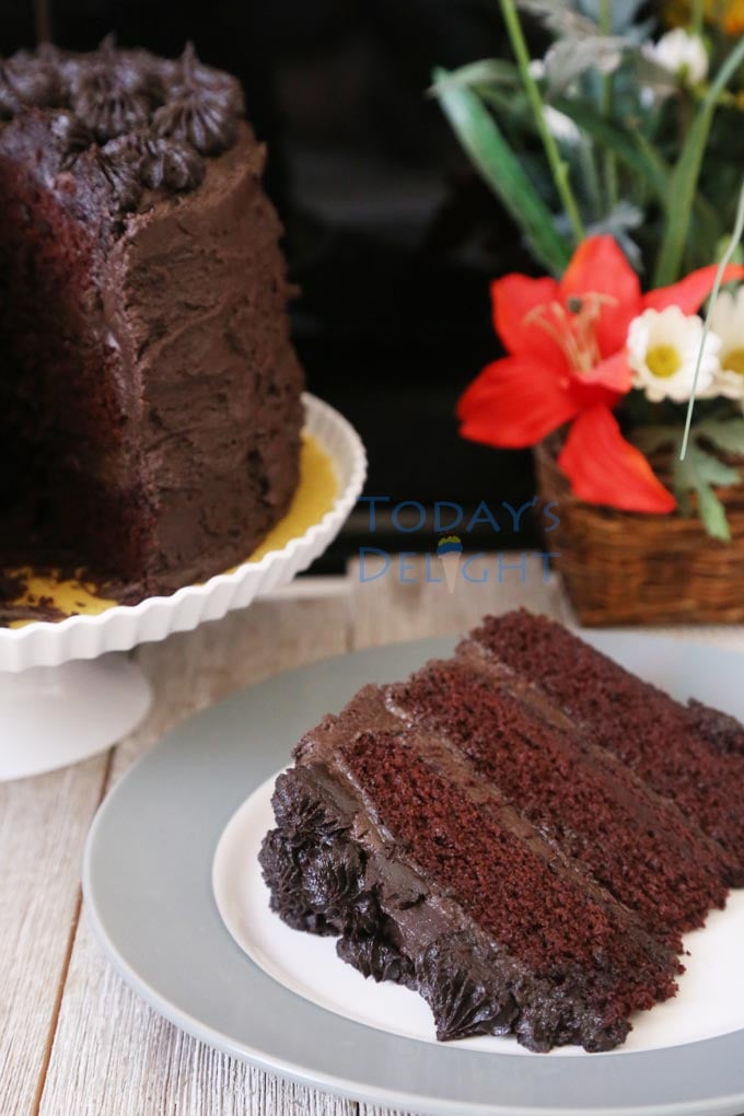 A slice taken from chocolate cake with dark chocolate cream cheese frosting on a cake stand and a slice of cake on a plate and a flower basket in the background.