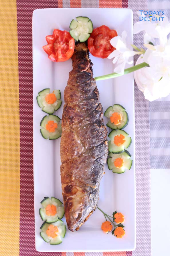 A whole Filipino rellenong bangus (stuffed milkfish) with decorative tomatoes, cucumber, and carrots on a platter on a colorful table runner and flower vase.