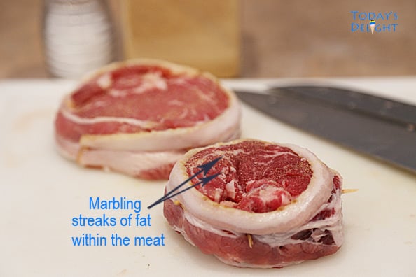 On this image I am showing marbling on two raw rib eye steaks wrapped in bacon. A salt shaker and knifes on a cutting board.