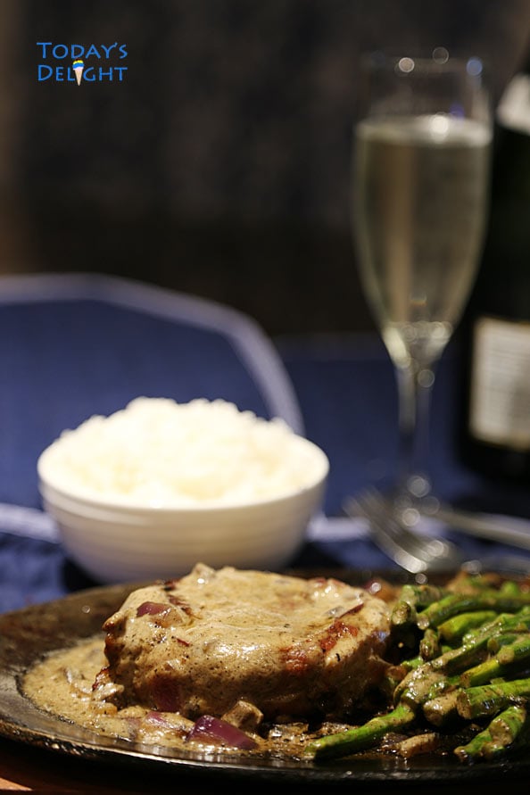 Rib eye steak wrapped in bacon with mushroom sauce and asparagus on a sizzling plate, a side of white rice and a glass of wine.