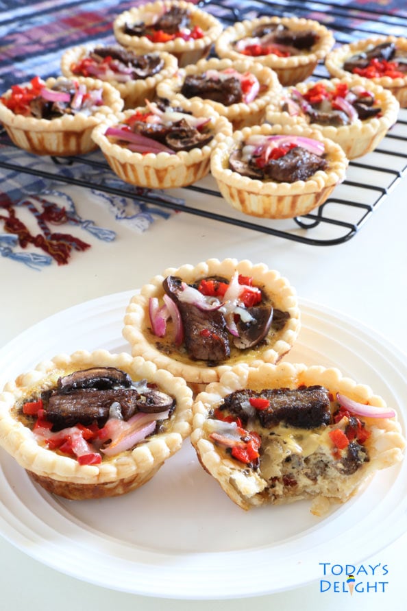 Cooling steak mini quiches topped with mushroom, onion and red bell pepper on a wire rack and a colorful kitchen towel nearby. Three mini quiches on a plate and one with a bite.
