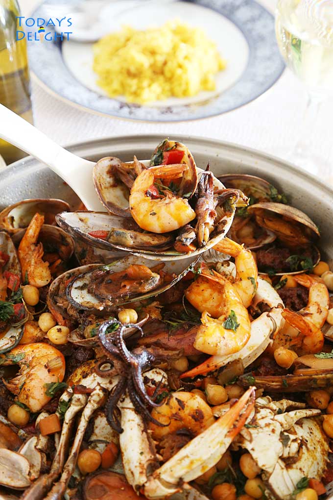 Spanish seafood stew with yellow rice, mixture of seafood, spicy chorizo, and vegetables in a cooking pan. Ladled stew to a plate.