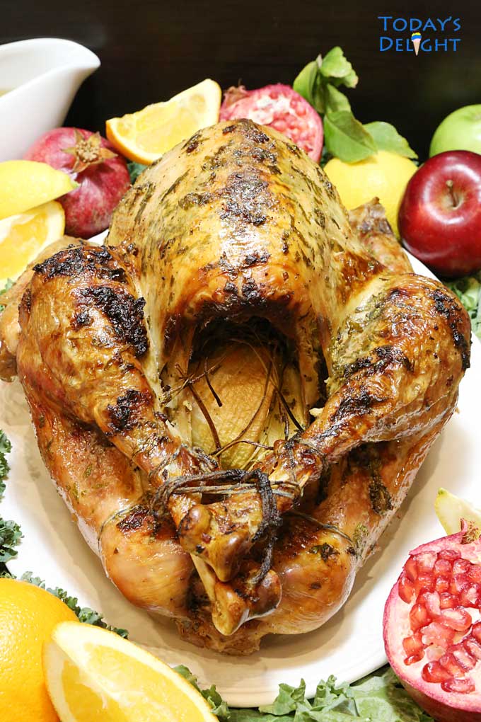 Roasted turkey's showing backend and surrounded with apples, pomegranates, lemons, kale and a sauce on the side.