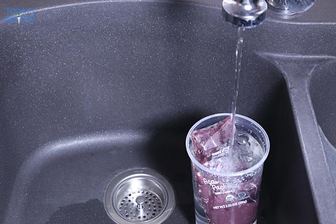 Acai puree packet is thawed in a glass over running cold water on the sink.