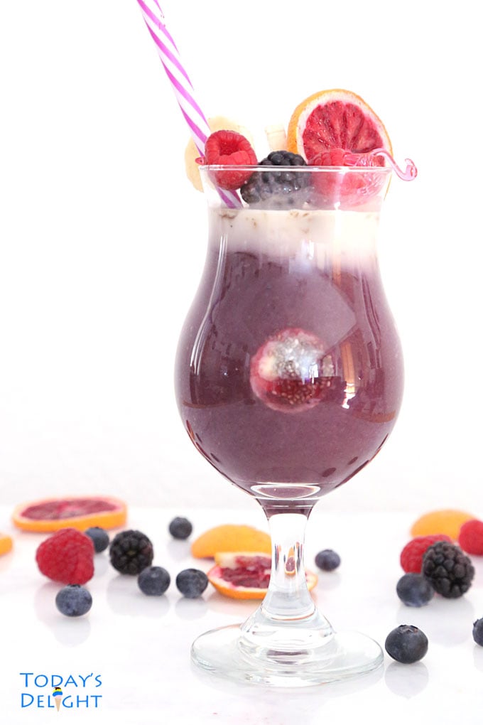 Coconut acai berry smoothie topped with raspberry, blackberry, decorative straw and a thin slice of blood orange on the glass. On the table are scattered blackberries, raspberries, and slices of blood orange.