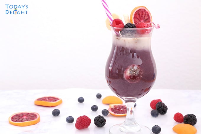 Coconut acai berry smoothie topped with raspberry, blackberry, decorative straw and a thin slice of blood orange on the glass. On the table are scattered blackberries, raspberries, and slices of blood orange.