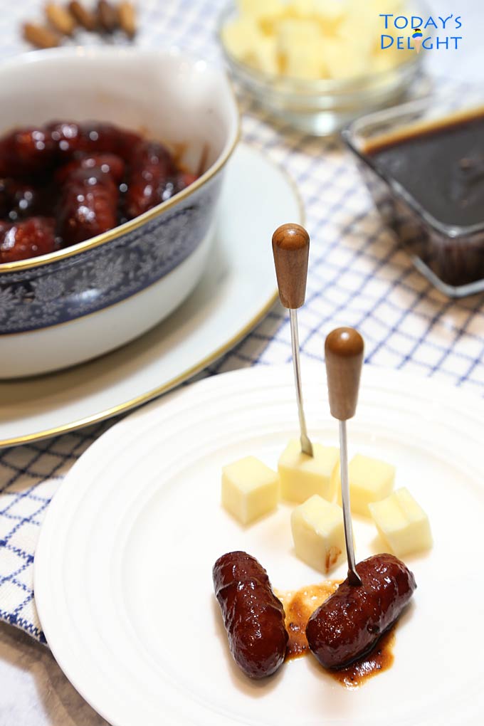 A couple of little smokies with one on an appetizer pick and another pick with cheese on a plate. In the background are bowl of lil smokies sauce, cheese and more appetizer picks.