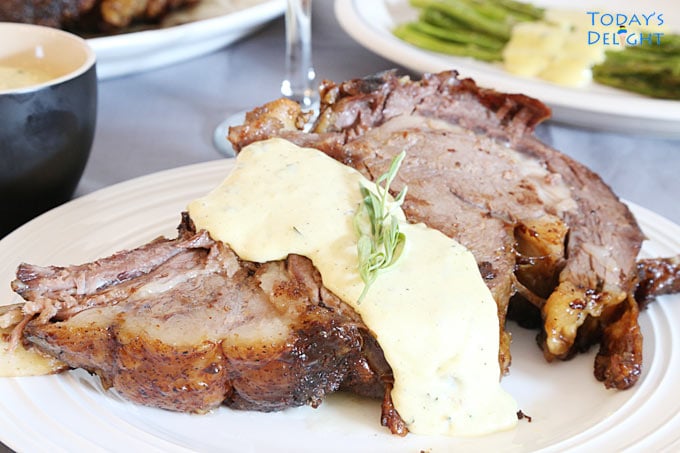 Two plates with steak with bearnaise sauce garnished with a sprig of tarragon, a side of asparagus, bearnaise sauce in a small bowl and a glass of red wine.