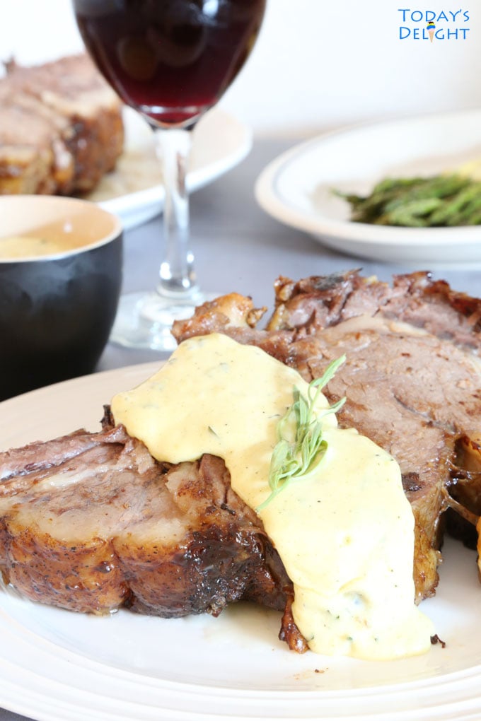 Two plates with steak with bearnaise sauce garnished with a sprig of tarragon, a side of asparagus, bearnaise sauce in a small bowl and a glass of red wine.
