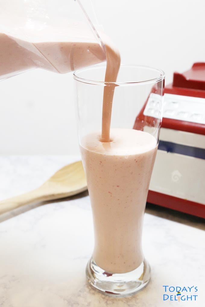Banana strawberry smoothie poured in a tall glass. Background show blender and a wooden big spoon.