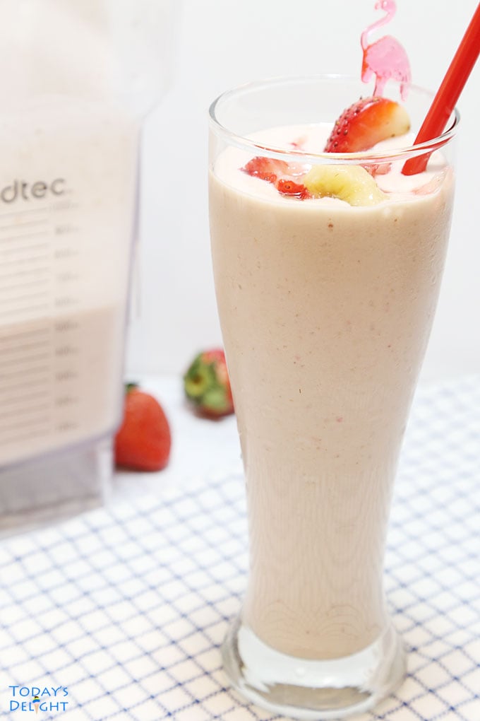 Strawberry banana smoothie garnished with sliced banana and a slice of strawberry with red straw. Background shows two strawberries and blender with smoothie.